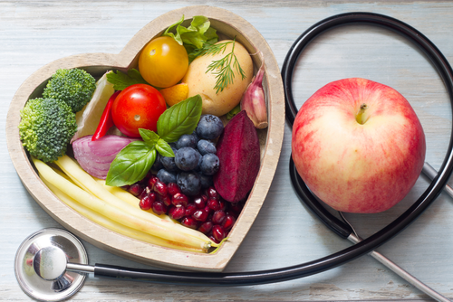 Fresh fruits and veggies in a heart shaped bowl next to an apple and stethoscope.