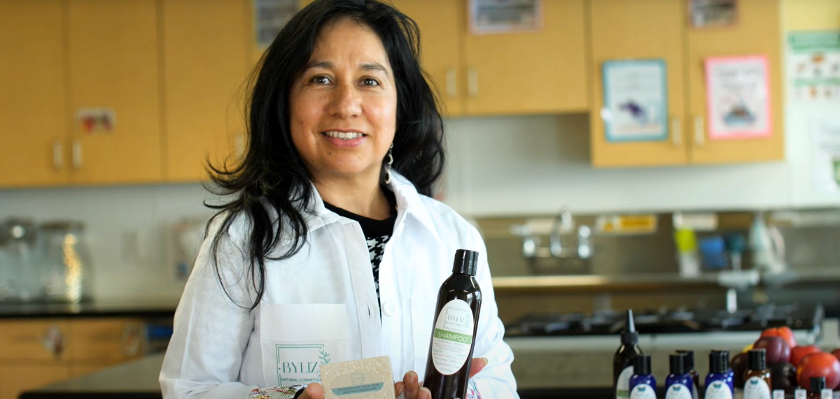 Elizabeth, wearing a white lab coat with her business's logo, holds up a soap and lotion.