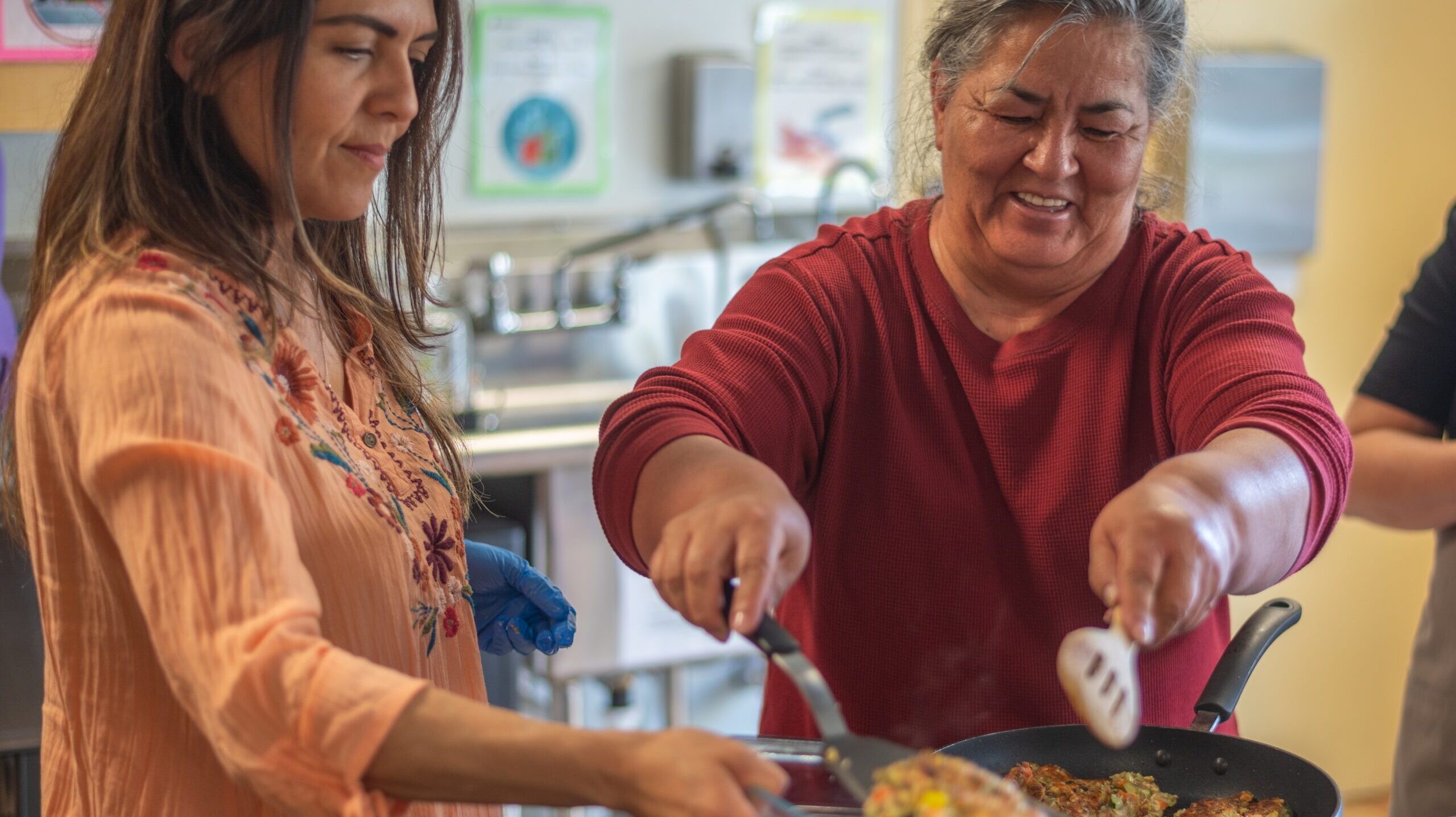 Two Latina women cook together in the Metro Caring kitchen. One holds a tray for the other. They're both smiling.