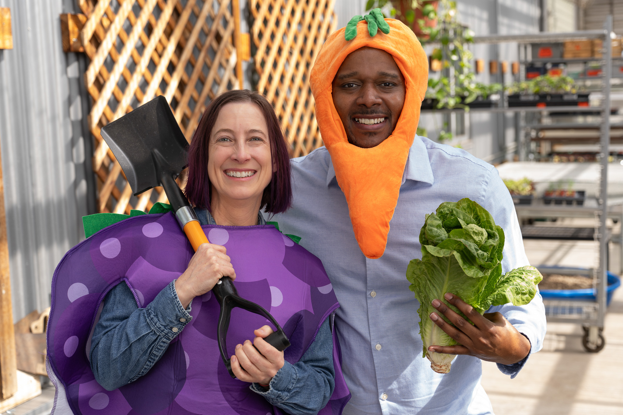 Teva, wearing a grape costume, holds a shovel over her shoulder, while Erik stands next to her with a carrot costume around his head while holding a head of lettuce.