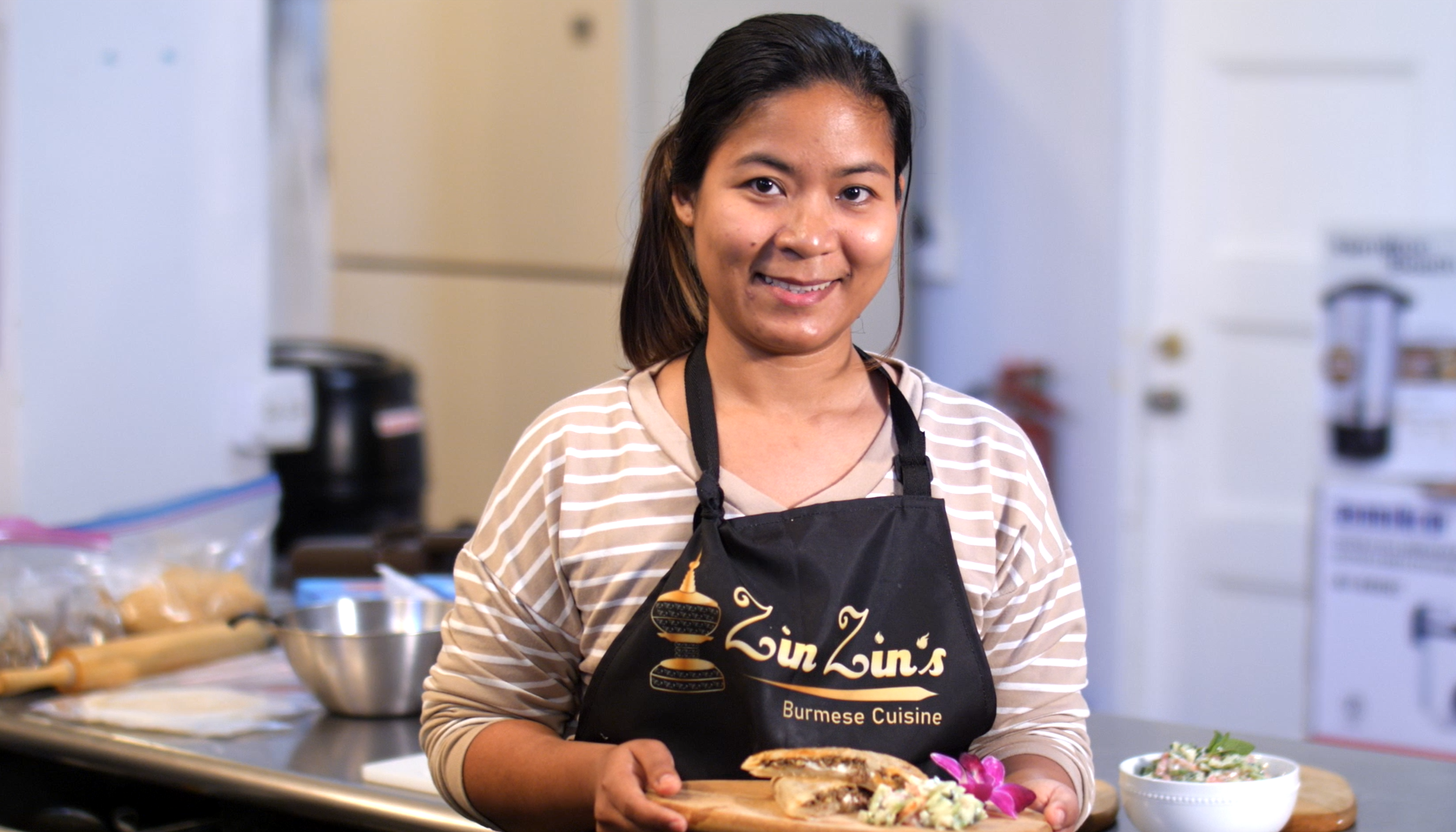 Zin Zin wearing her branded restaurant apron, holds up a plate of Burmese food.