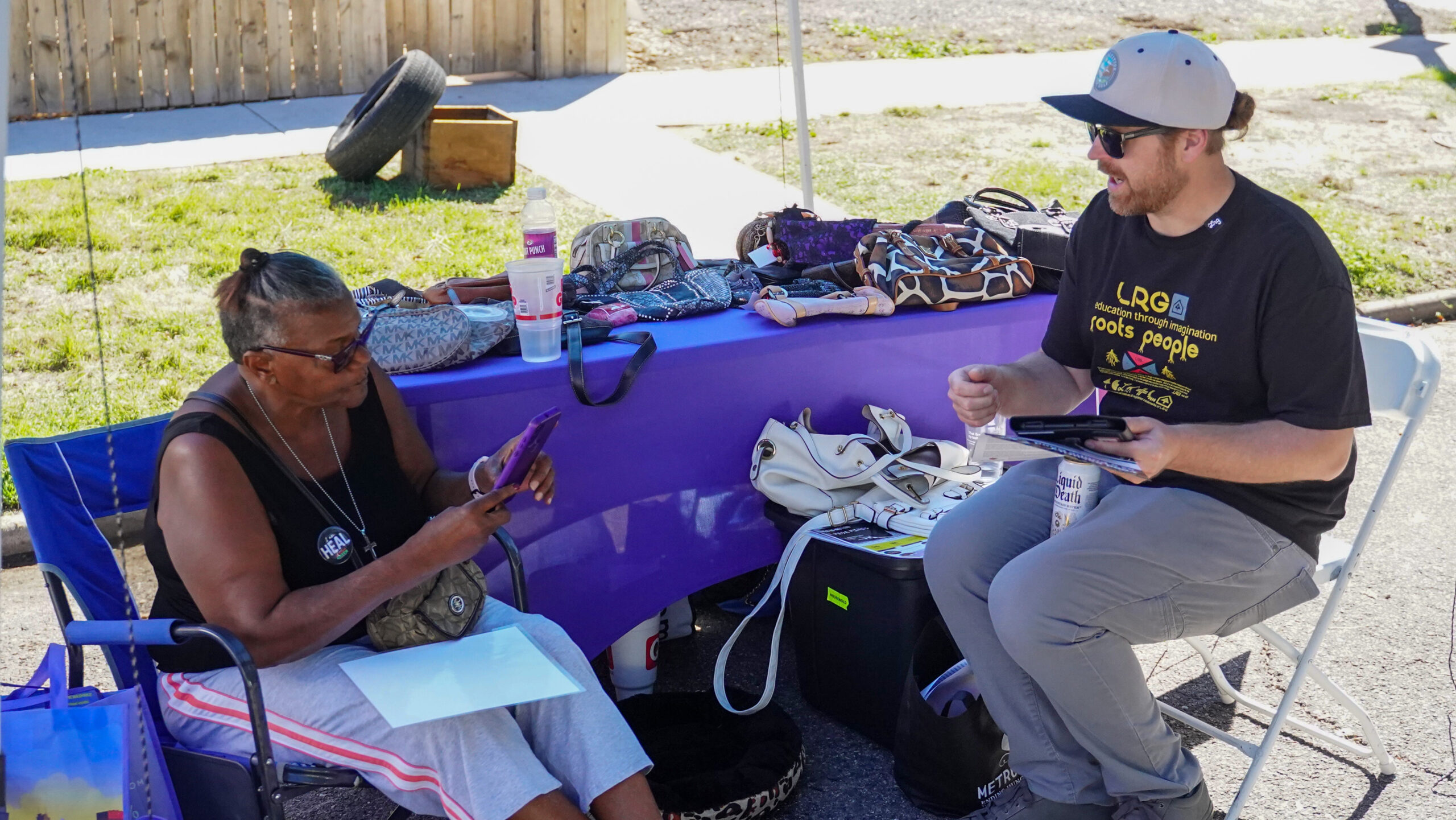 Shan and a community member sit in lawn chairs in front of a vendor's table.