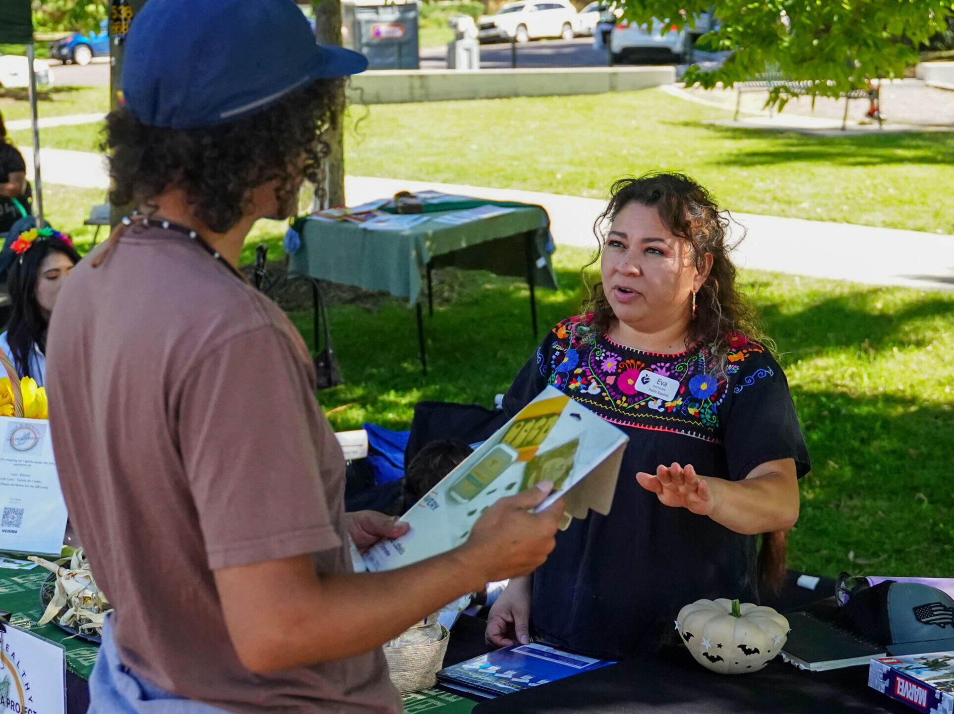 Two people chat at a table stand outside. One person is holding a clipboard.