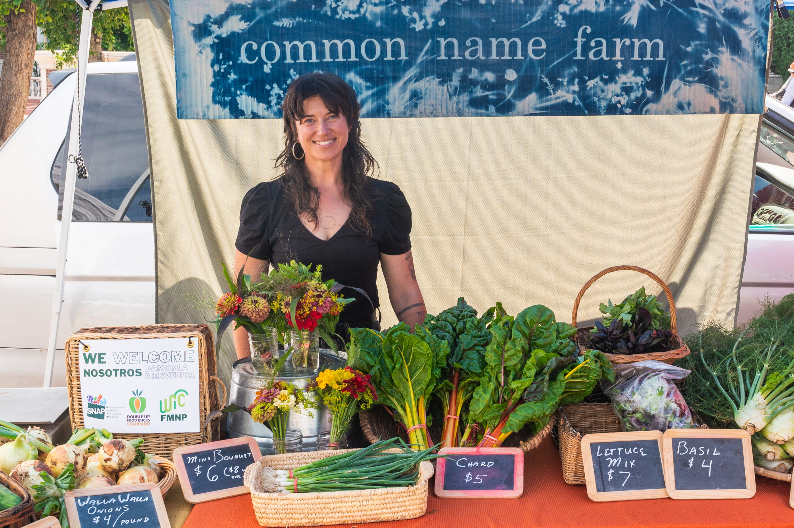 Betzi stands behind a table full of veggies for sale, including carrots, rainbow chard, lettuce, basil, and some flowers.