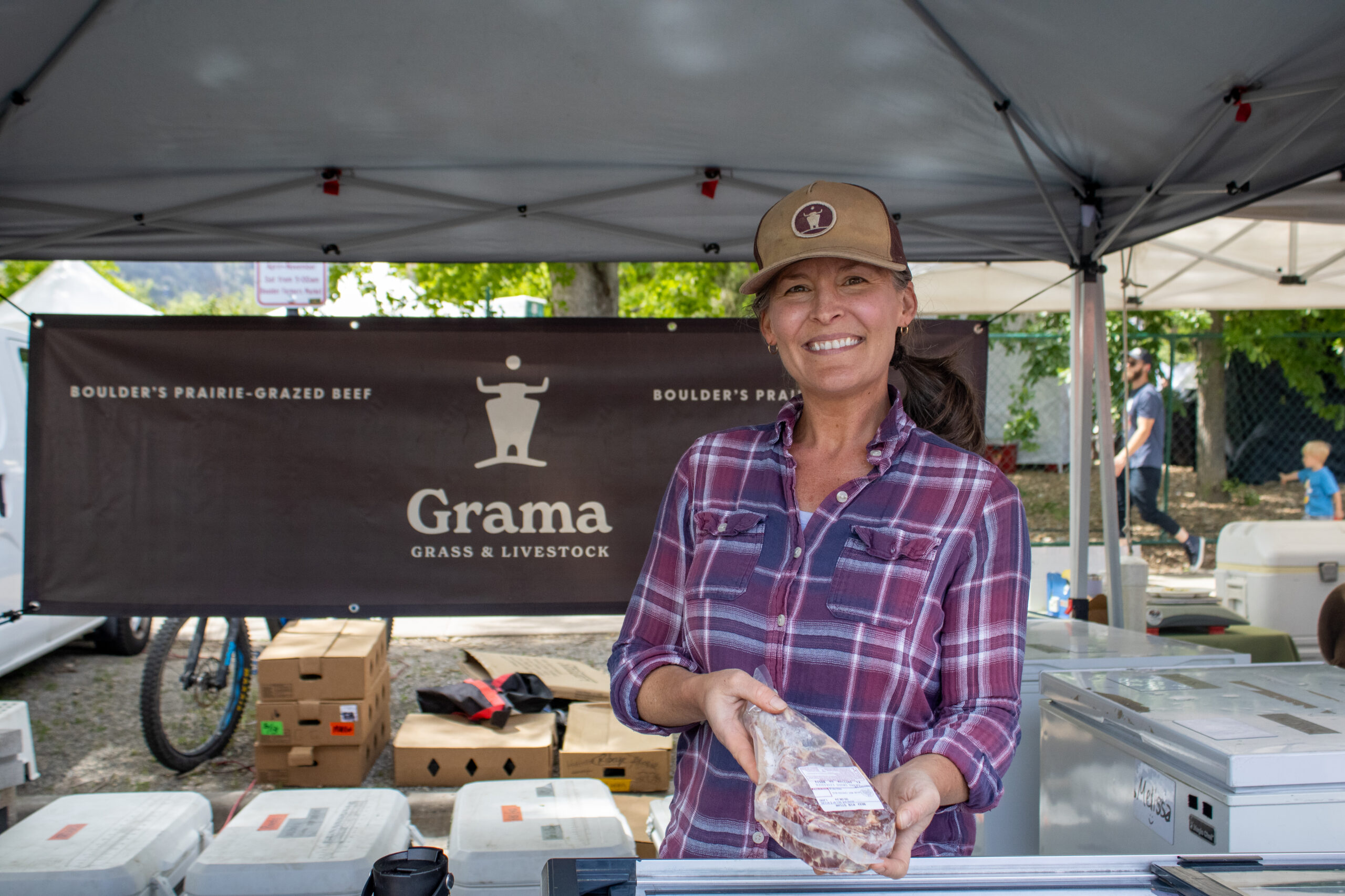 Julia Wolfe holds up a piece of wrapped beef behind the Grama Grass table at the farmers market. She's wearing a purple flannel and Grama Grass branded trucker hat.