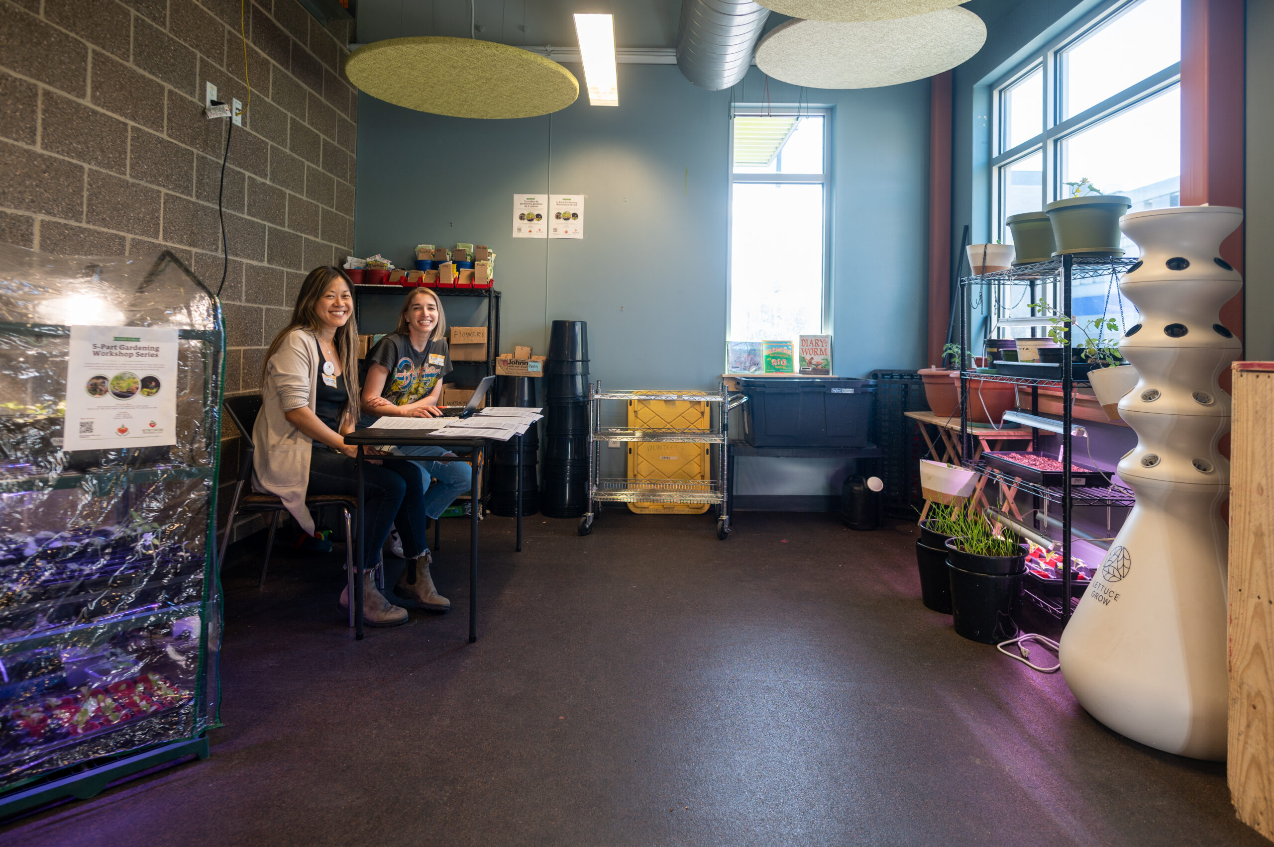 Two volunteers sit in the Indoor Garden smiling at the camera. In view is multiple setups of plants on shelves with grow lights shining on them. 