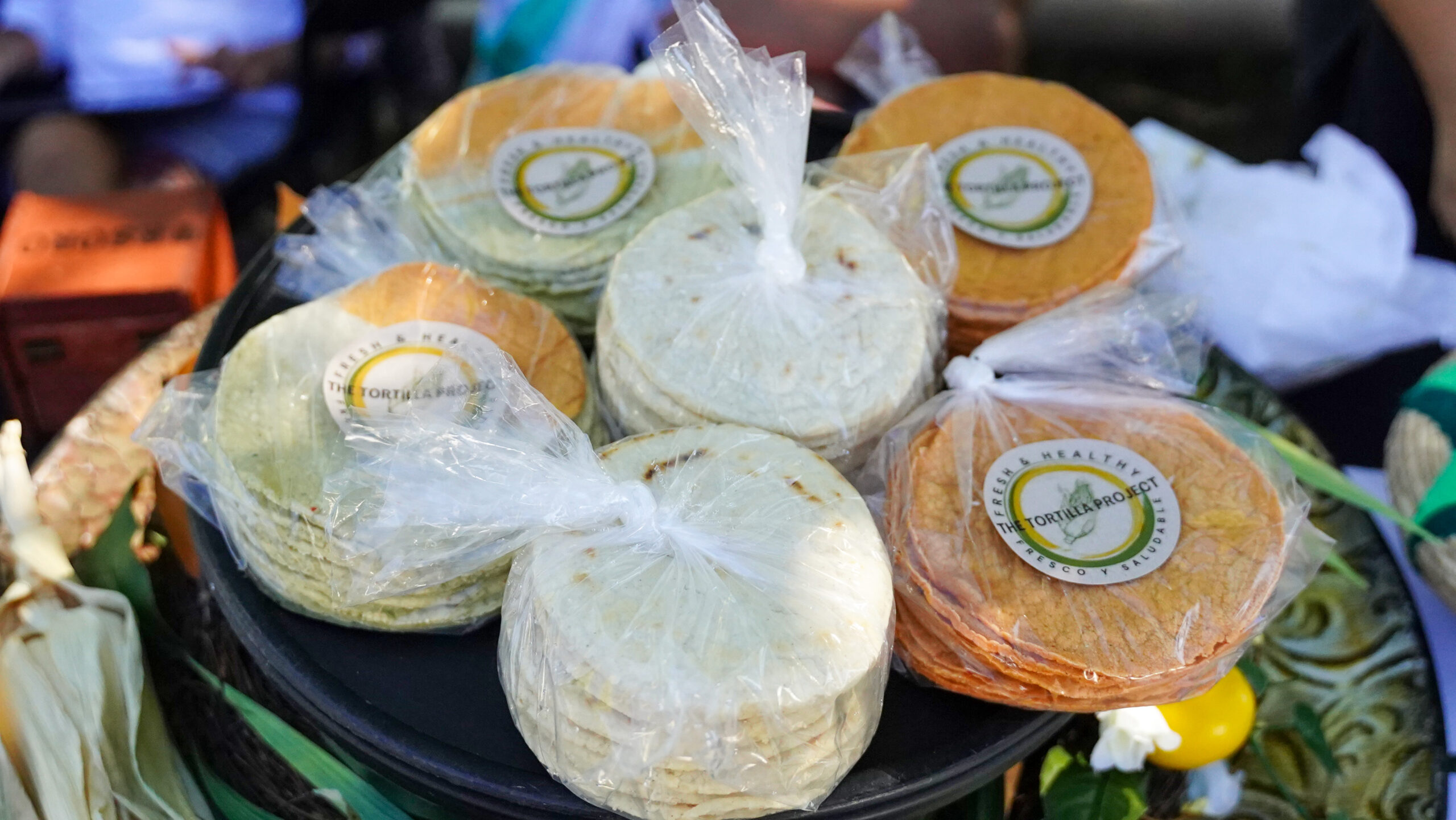 Close up of tortillas of orange and tan colors, wrapped in plastic, with a sticker for The Tortilla Project.