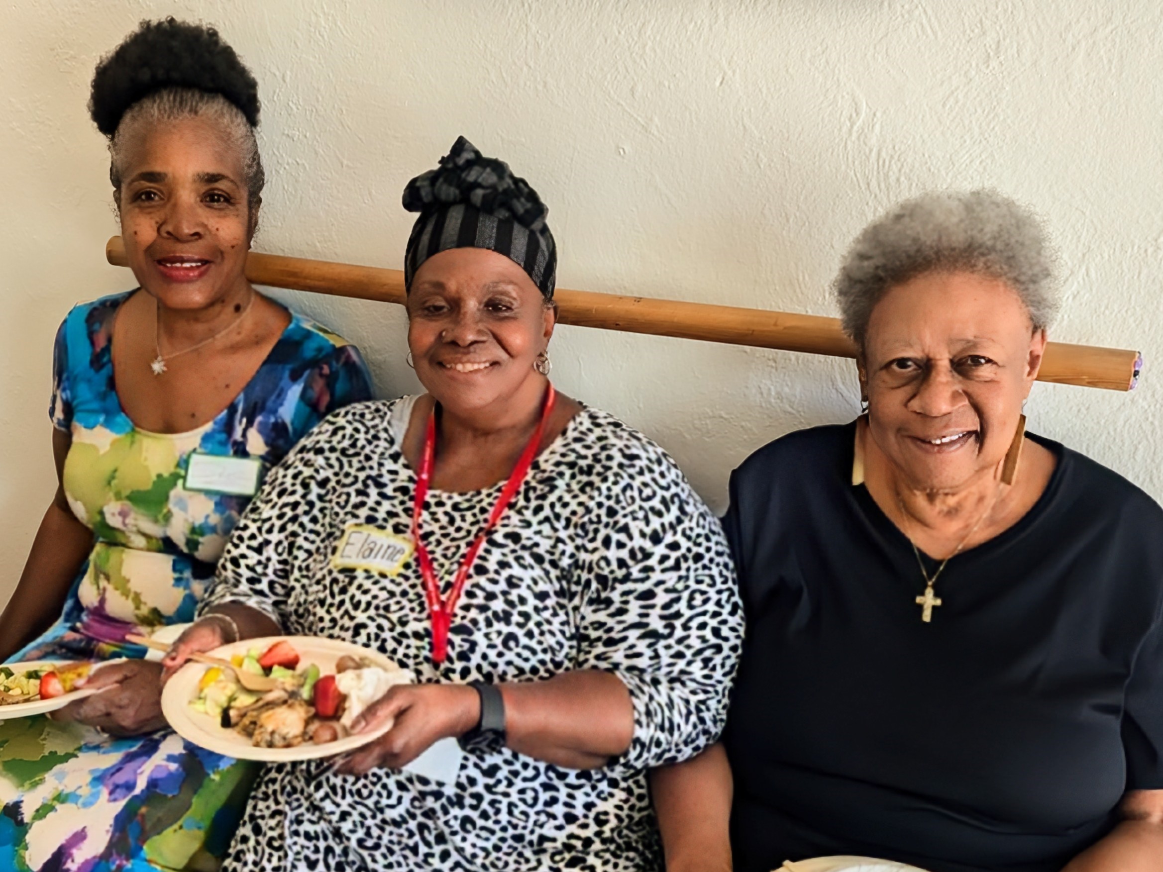 Three women sit together eating food and smiling at the camera. 