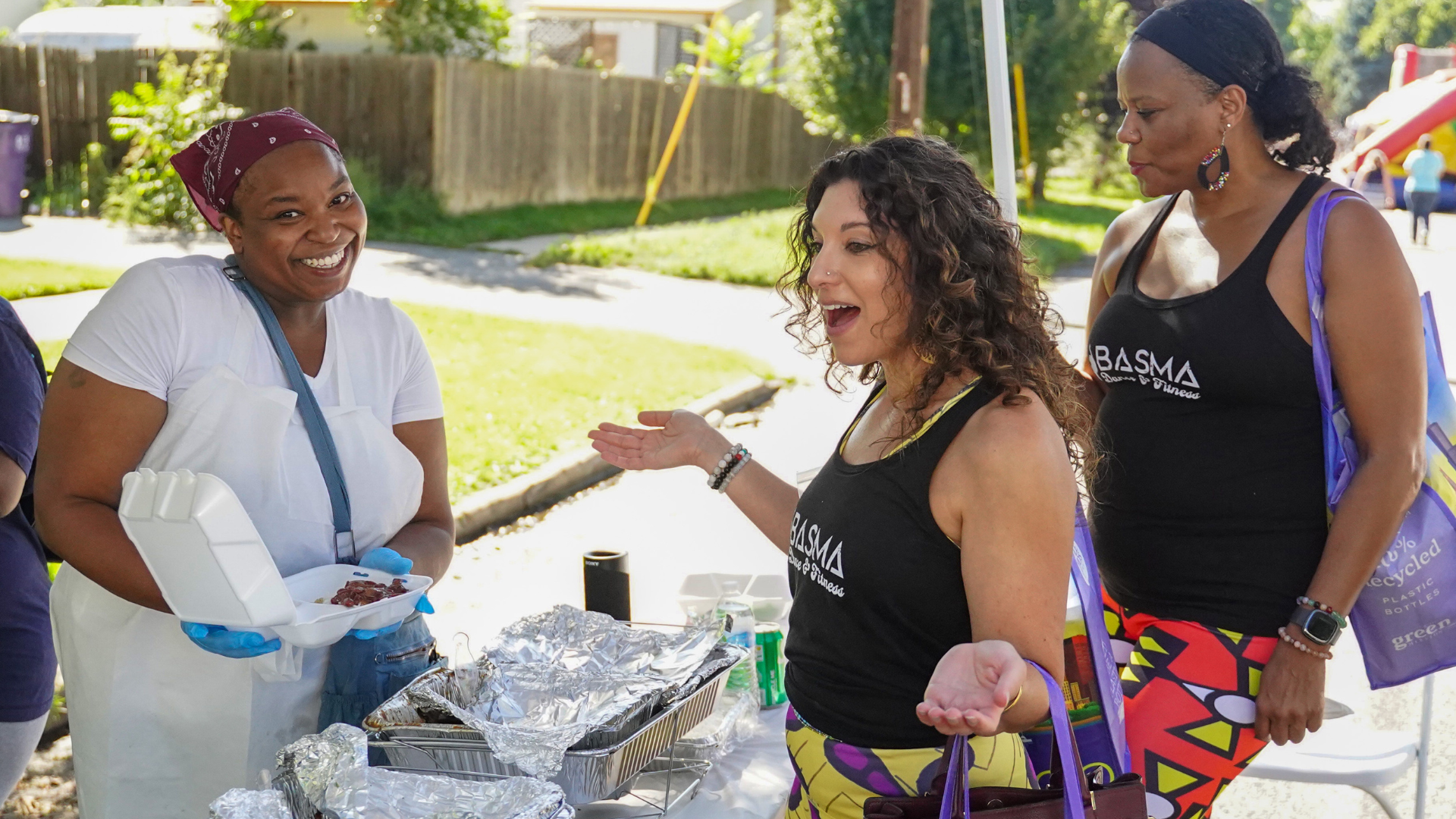 Two people approach an outdoor food vendor, who is holding up a to go container of food, smiling. One attendee smiles widely with her hands up.