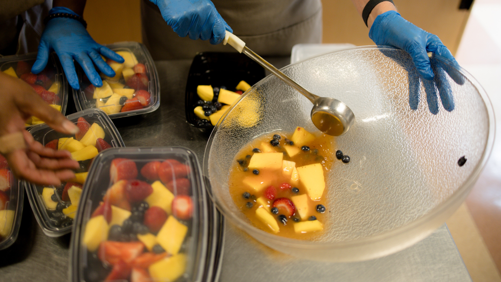 A big bowl of mixed fruit is being scooped into smaller prepped containers of fruit salad. People's hands (with gloves) are in the frame.