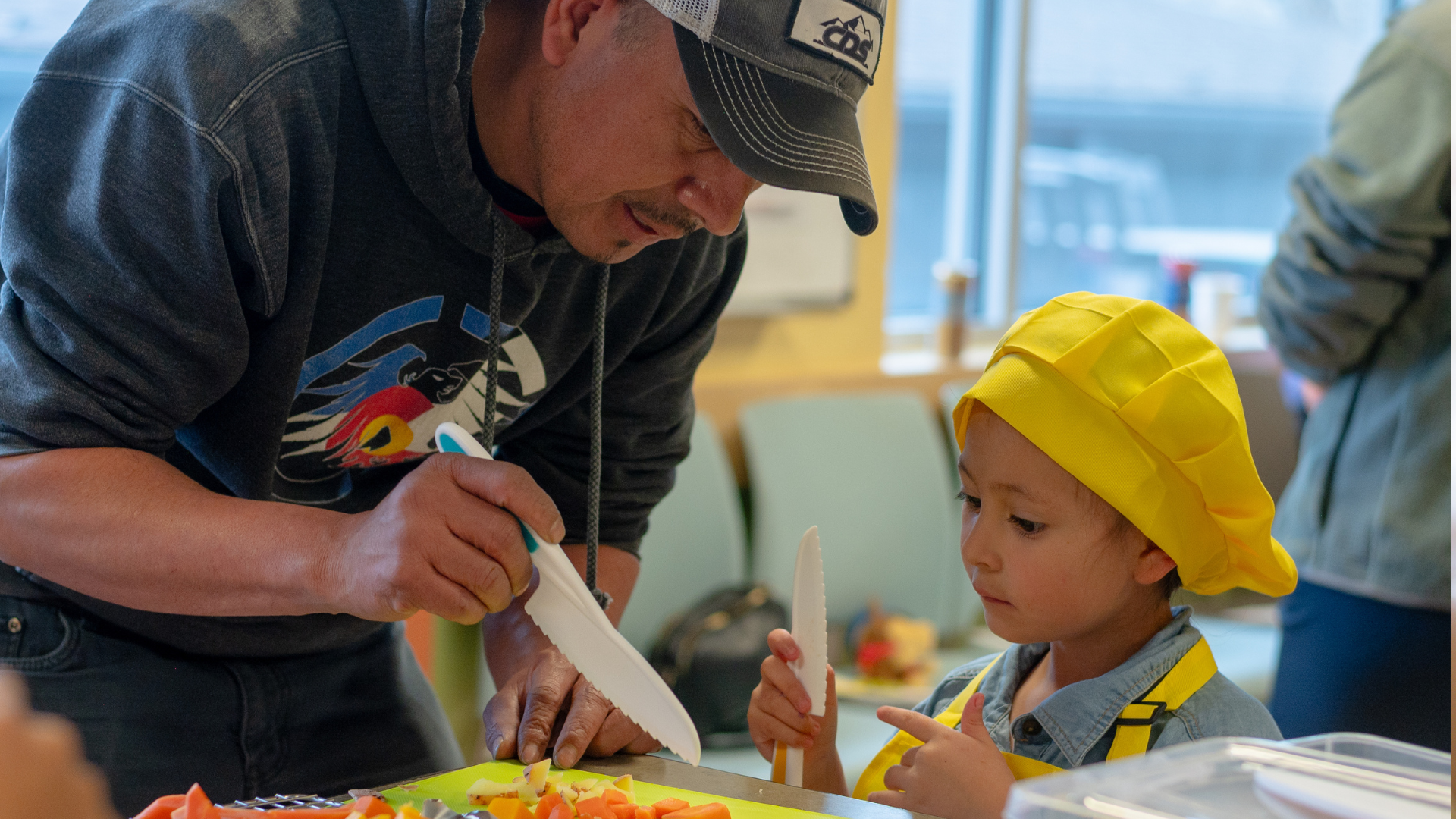 A dad is explaining something to his child about the veggies they are cutting. The child is wearing a bright yellow chef's hat.