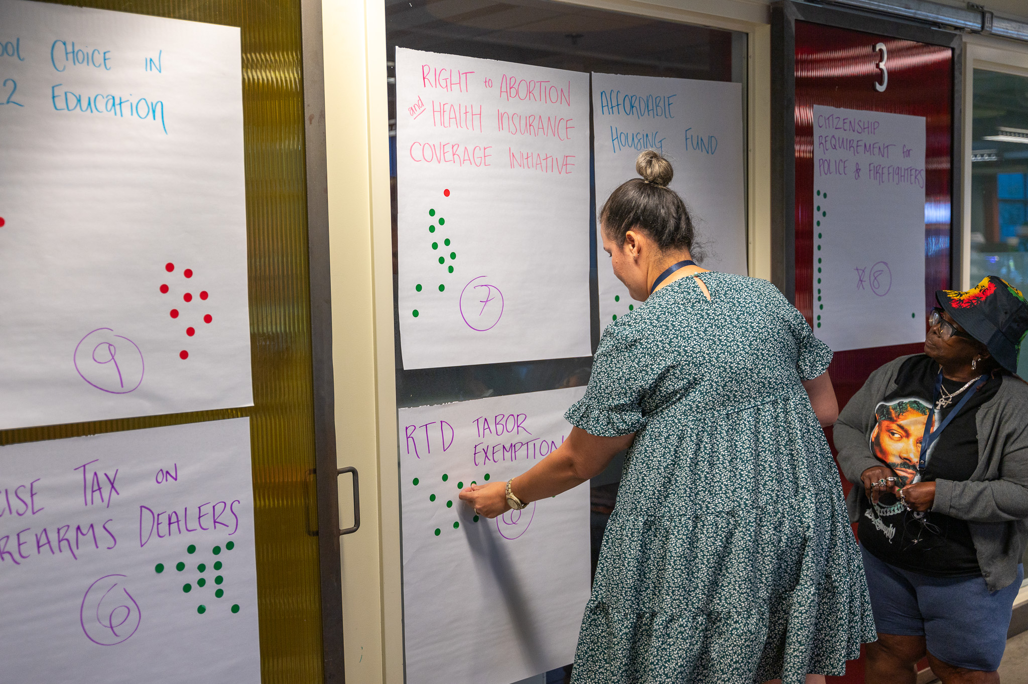 Community member adds a sticker to a large white sheet of paper stuck to the wall next to other sheets of paper that each have a different ballot initiative on them. 