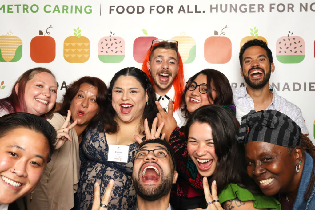 Ten staff, volunteer, and community members take a silly photo with huge smiles in a photobooth with a background that reads "Food for all. Hunger for none." 