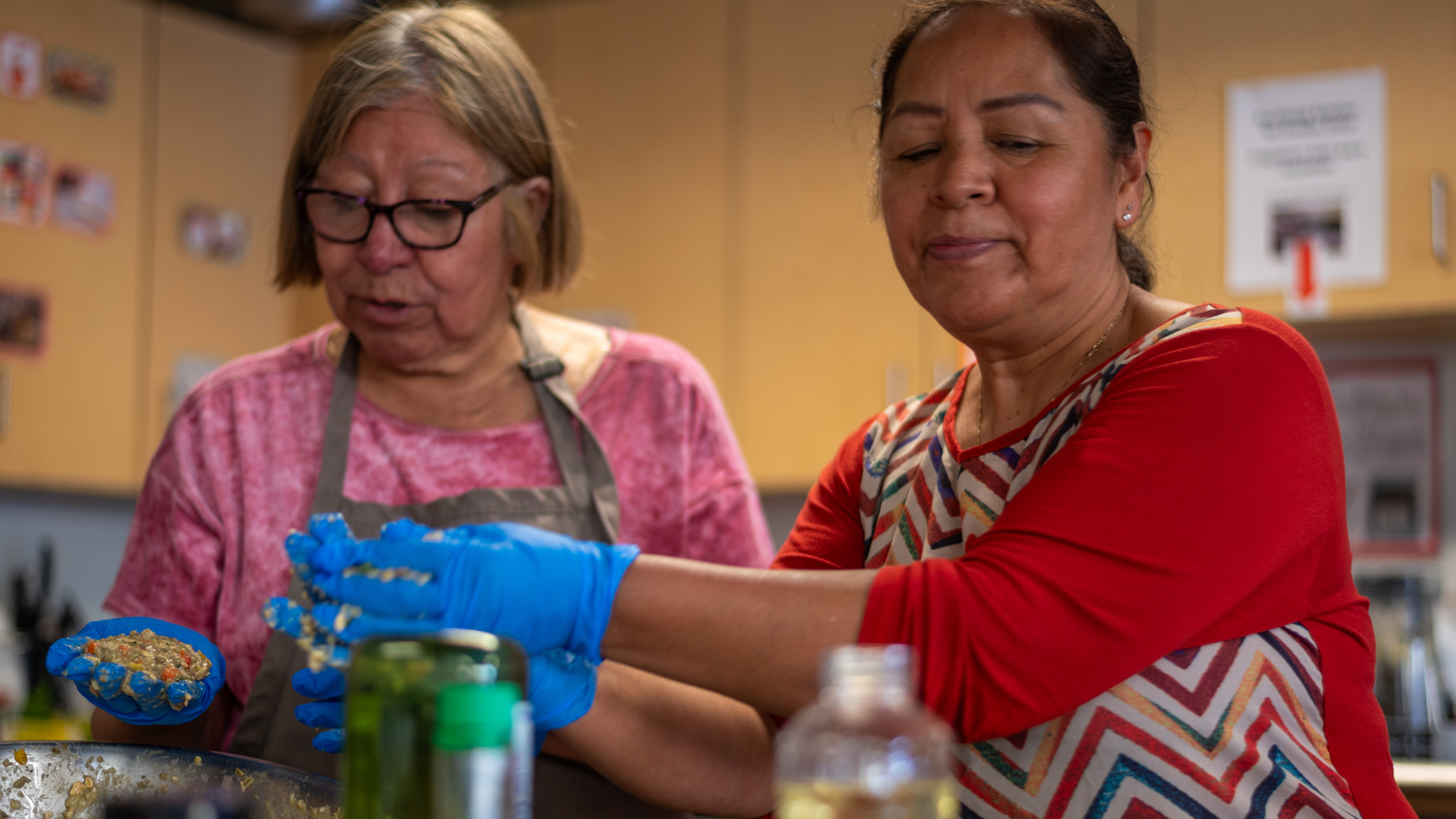 Two women cook together in the Metro Caring kitchen. One, while wearing gloves is moving a patty to the griddle.