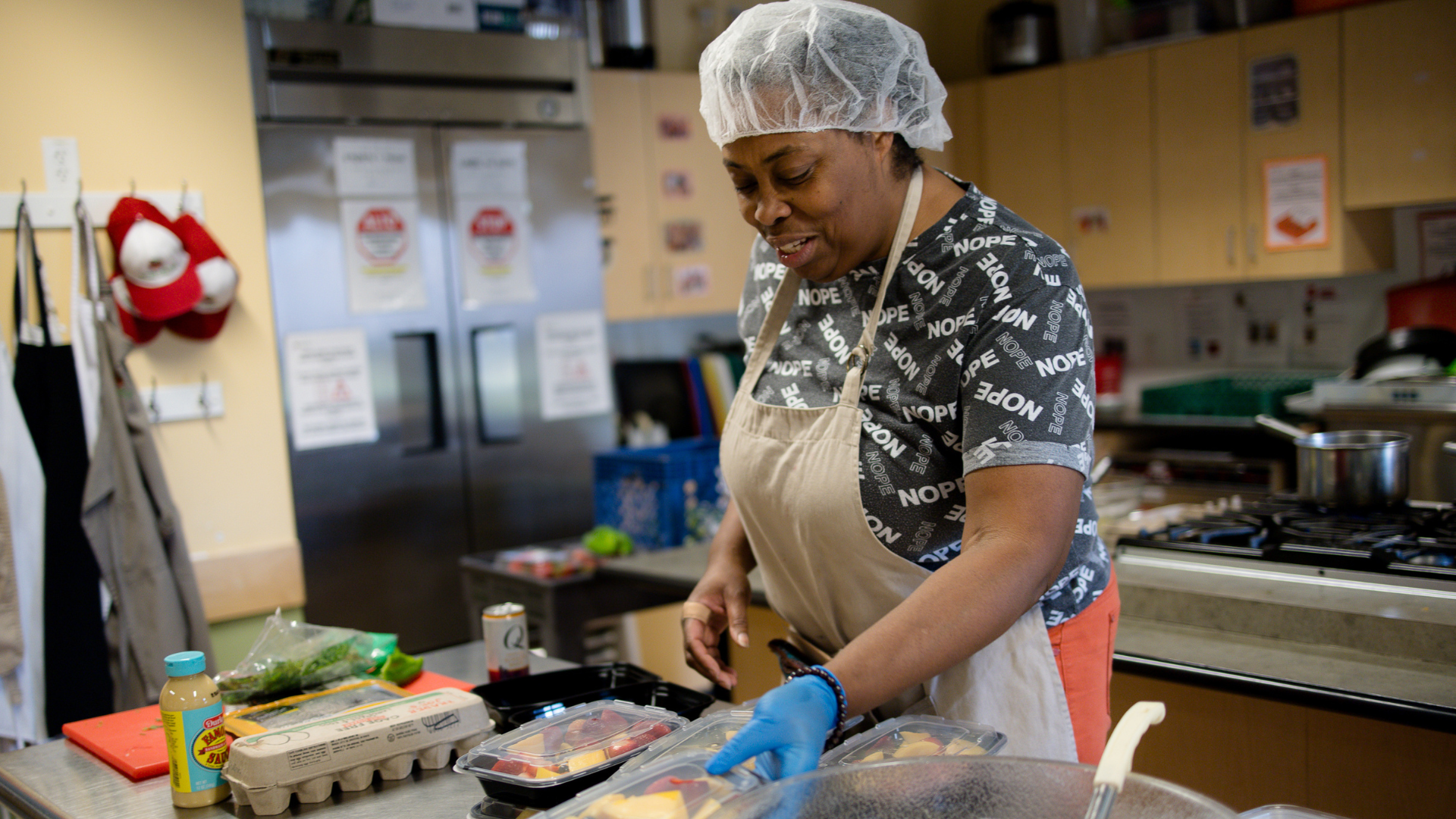 Chef Ronnie prepares fruit salad containers in the Metro Caring kitchen. She's smiling while wearing a hairnet, gloves, and apron.