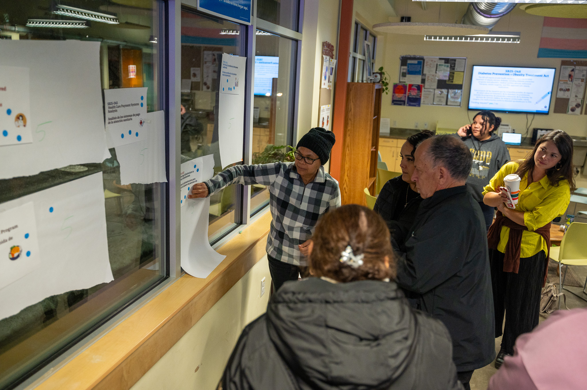 Six community members gather around the windows that have 10 pieces of paper taped up listing different bills. 