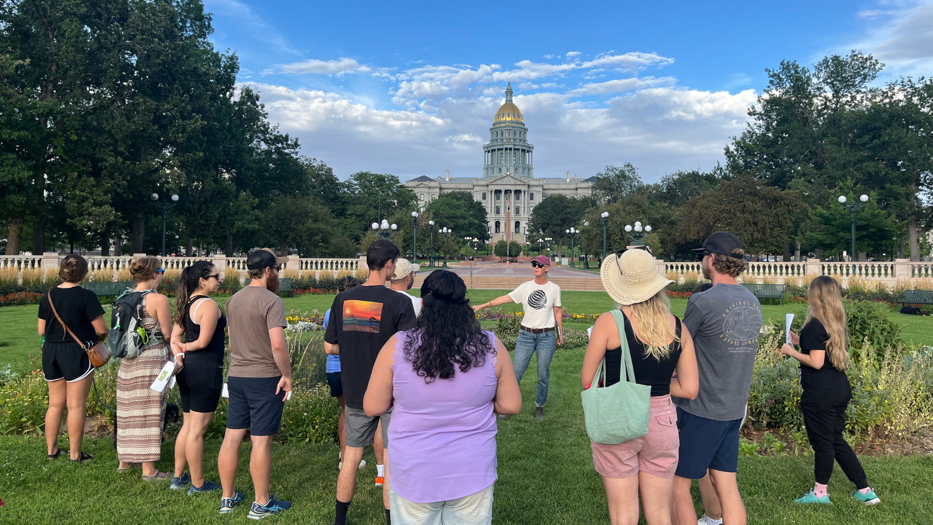 Group of people listen to Linda from Grow Local Colorado in the gardens. The Colorado Capitol building is in the background.