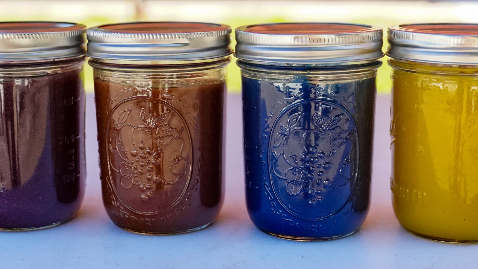 Close up of 4 mason jars filled with vibrant colors of preserved items: one is dark purple, one dark red, one blue, and one yellow.