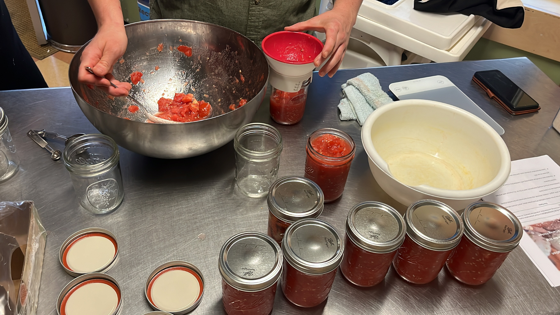 Overhead view of a person scooping chopped produce into a mason jar can. Several other filled jars line the table.