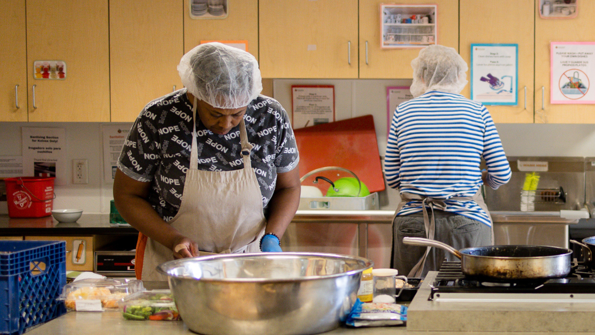 Two people cook in the Metro Caring kitchen. One is preparing ingredients on the prep counter while the other is doing dishes in the multi-compartment sink behind.