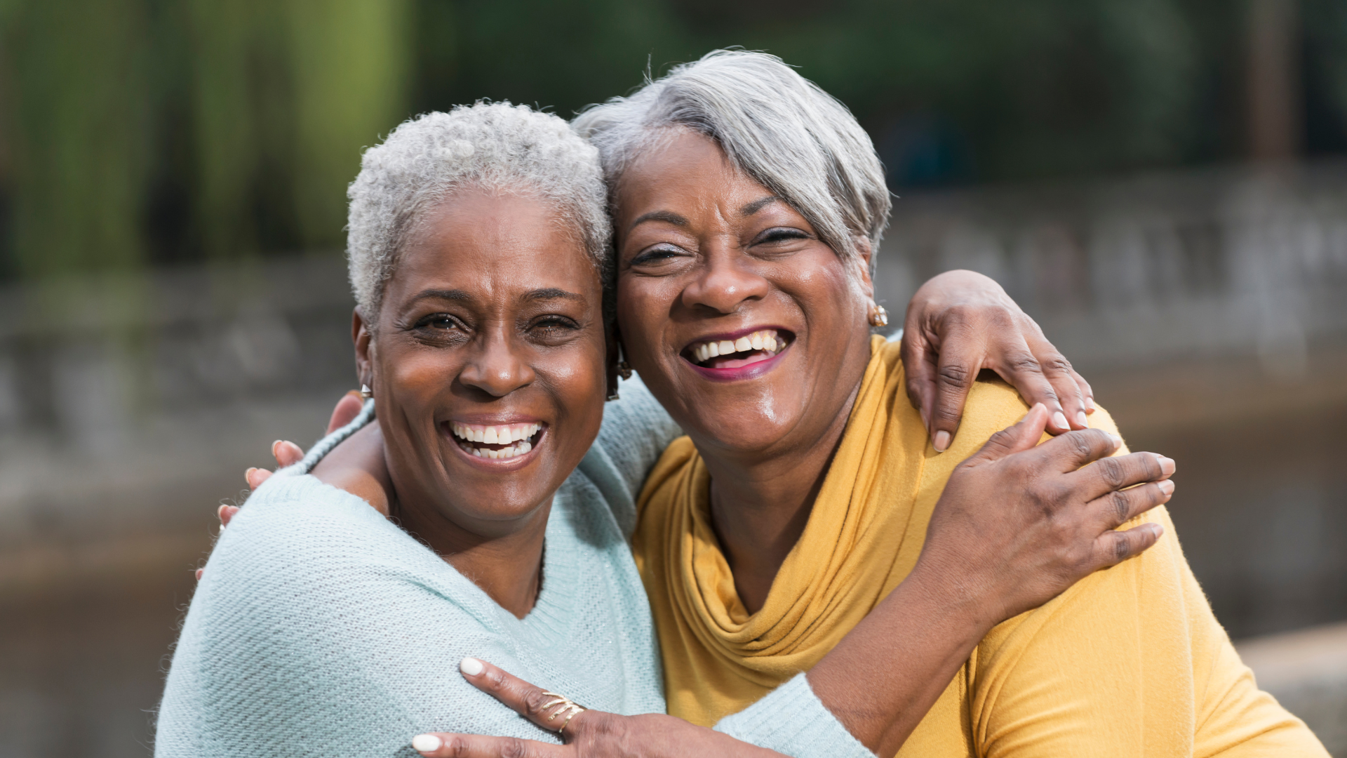 Two Black women with silver hair hug each other while smiling at the camera.