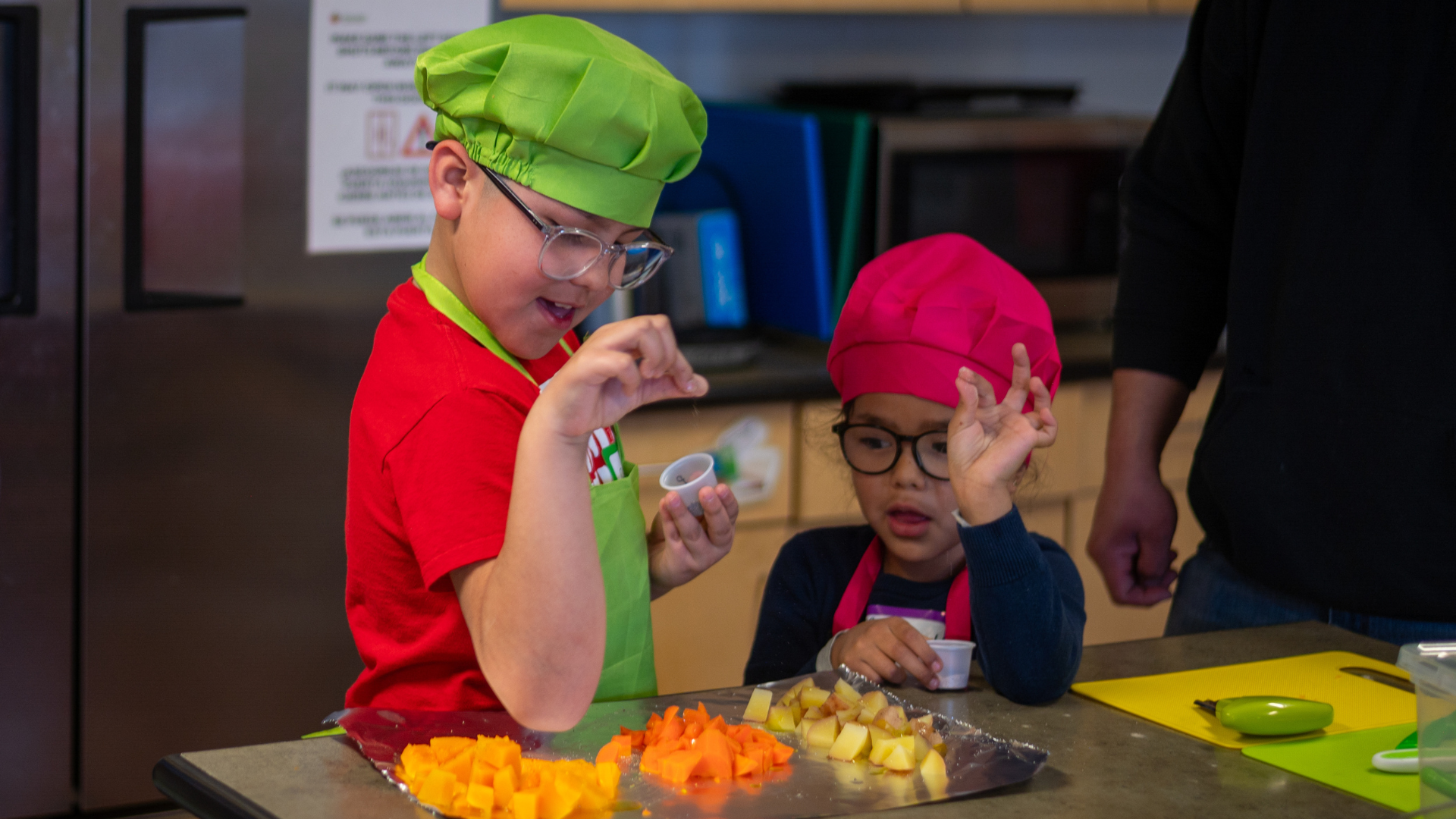 Two young children wearing colorful chef hats and aprons are seasoning chopped vegetables together in a kitchen setting.