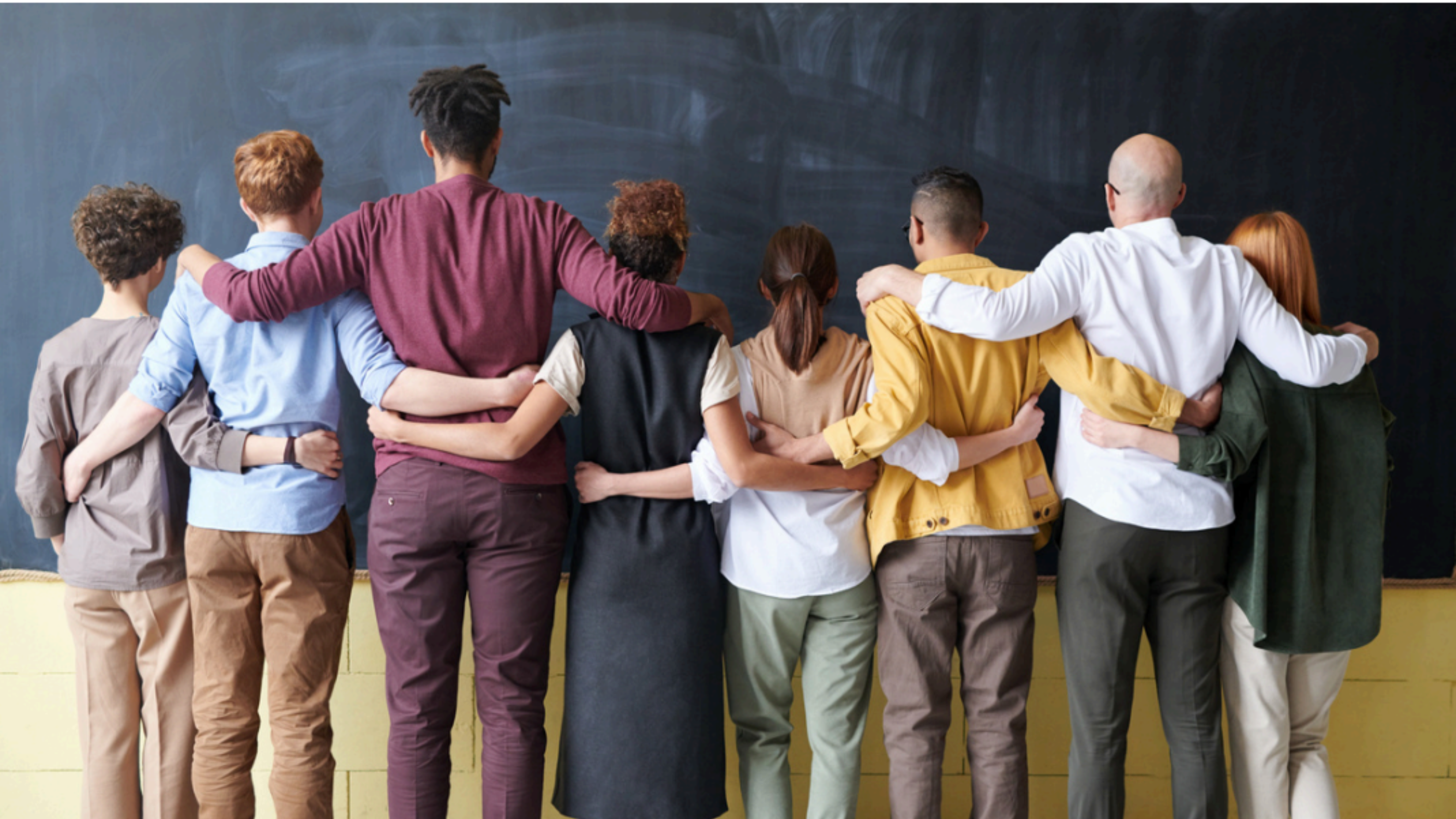A diverse group of nine people standing side by side with their arms around each other's backs, facing a blackboard. They are dressed in casual clothing, symbolizing unity, teamwork, and inclusivity.