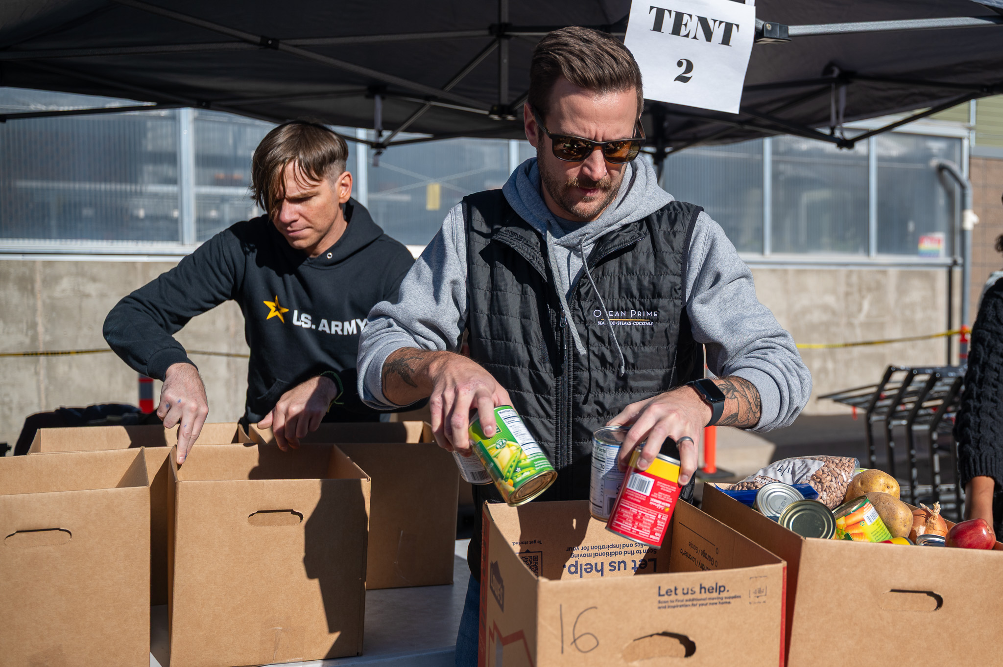 Two volunteers load cans into cardboard boxes sitting on a small shopping cart underneath a tent outside of Metro Caring. 
