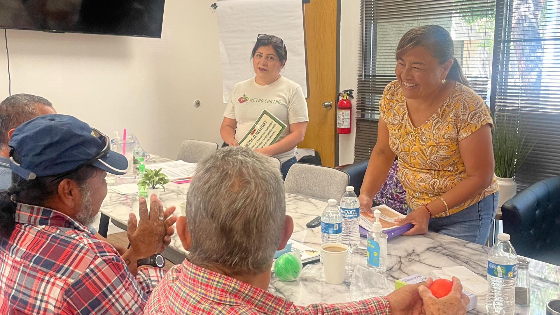 Latina woman holds a packet of papers, smiling, while she stands at a table talking to several people in Metro Caring's Diabetes Among Friends support group/