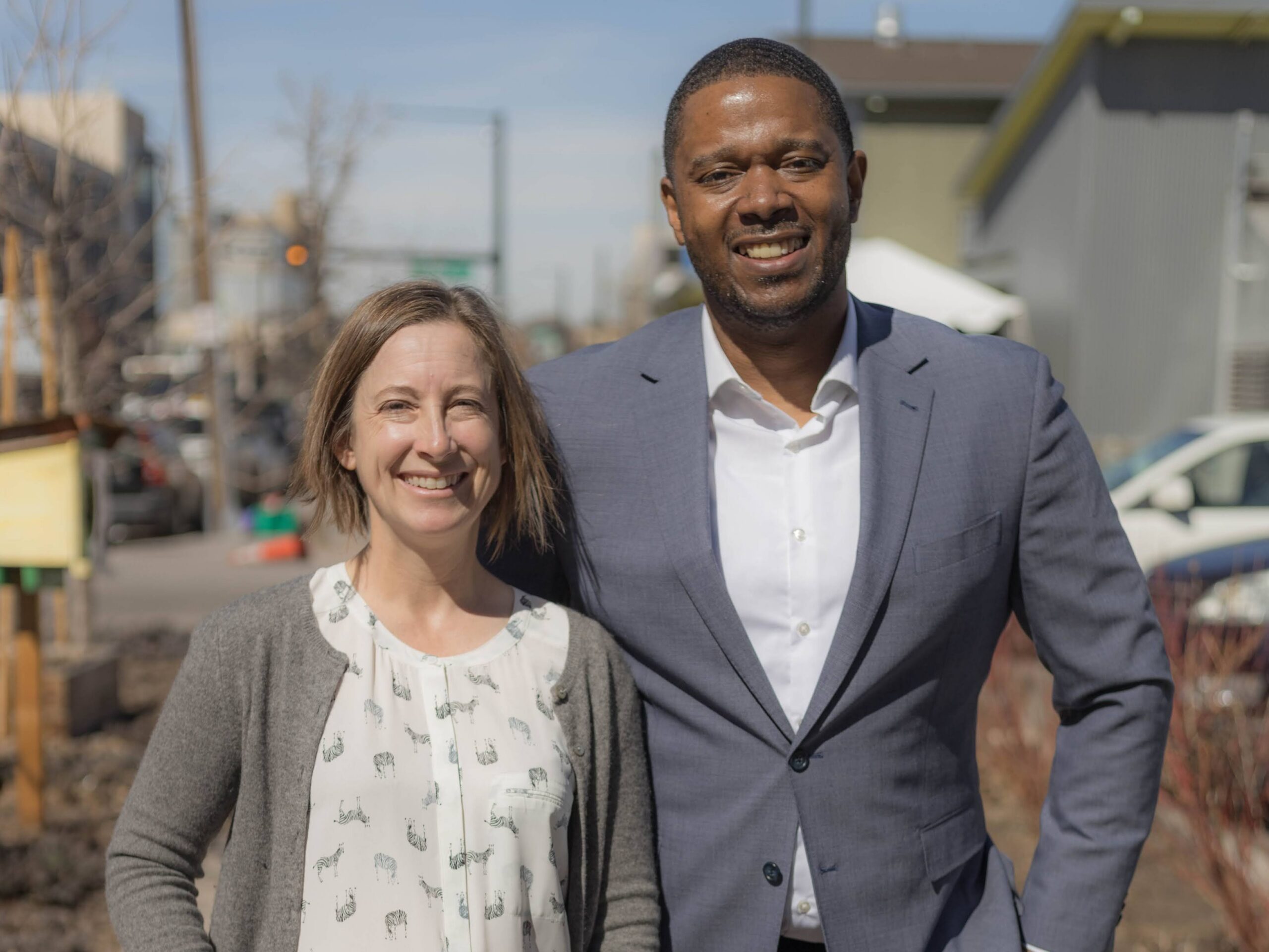 Erik and Teva stand shoulder to shoulder outside of Metro Caring's building, smiling at the camera.