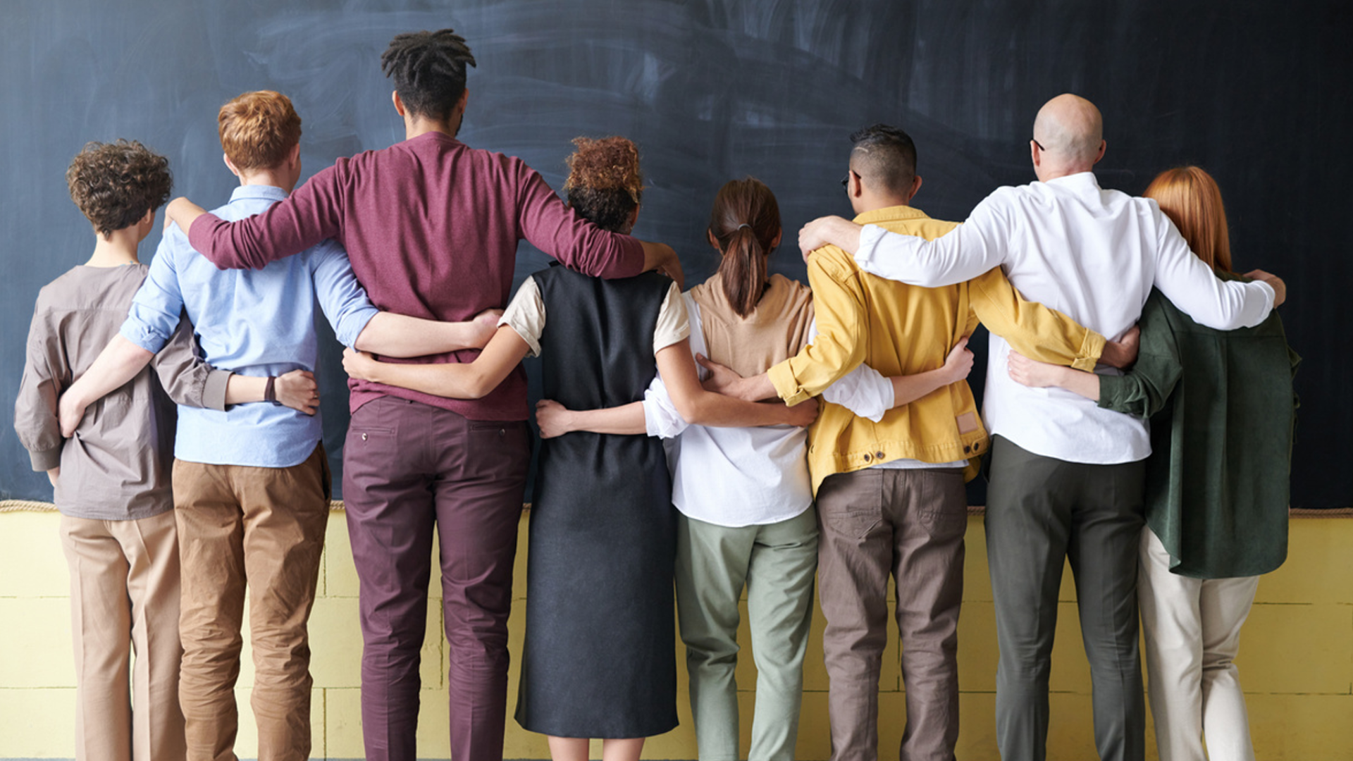 Eight people of all different races face a chalk board with their arms over each other's shoulders.