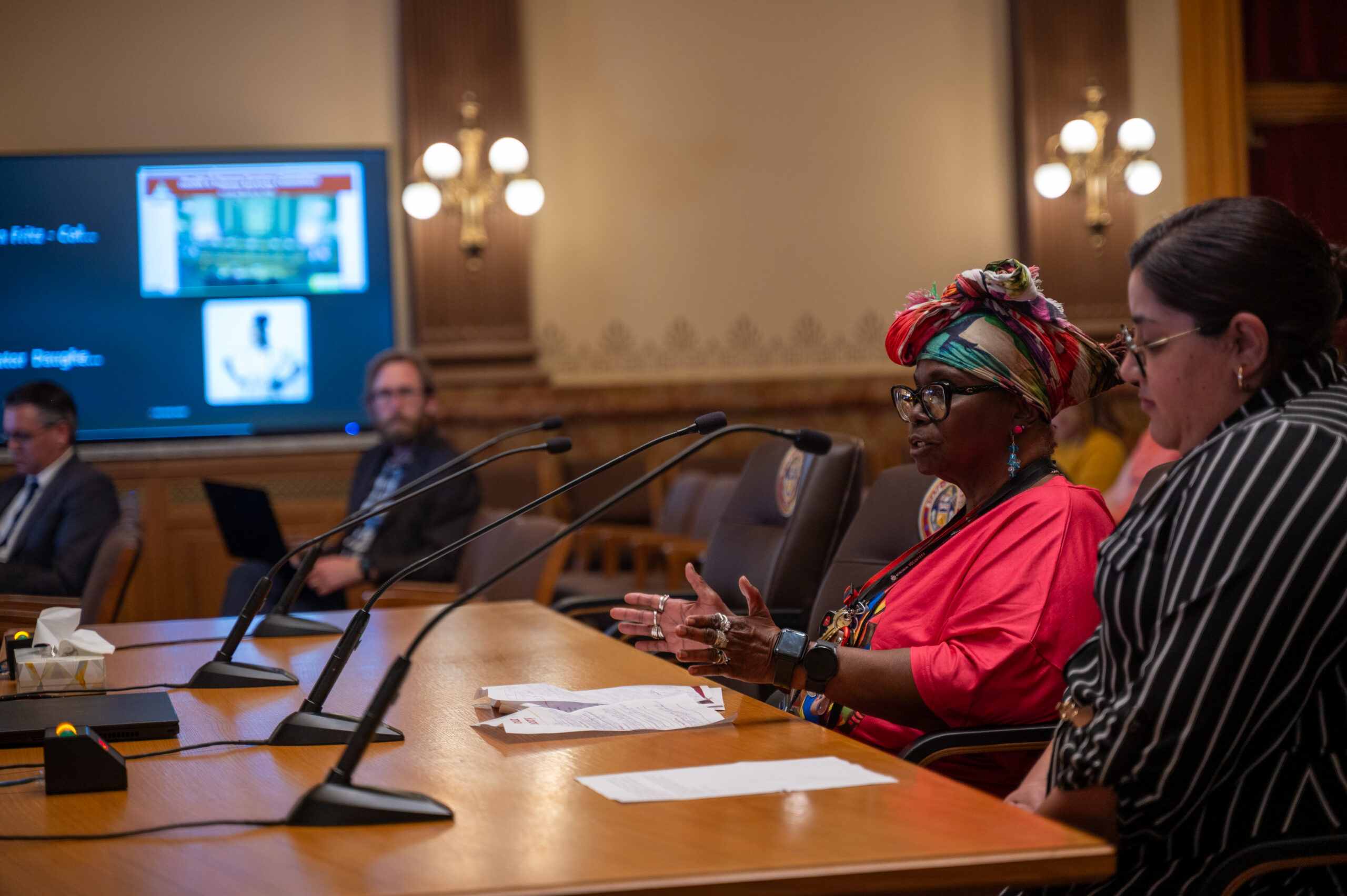 A Black woman speaks into the microphone while sitting at a long wooden table in the Capitol building. 