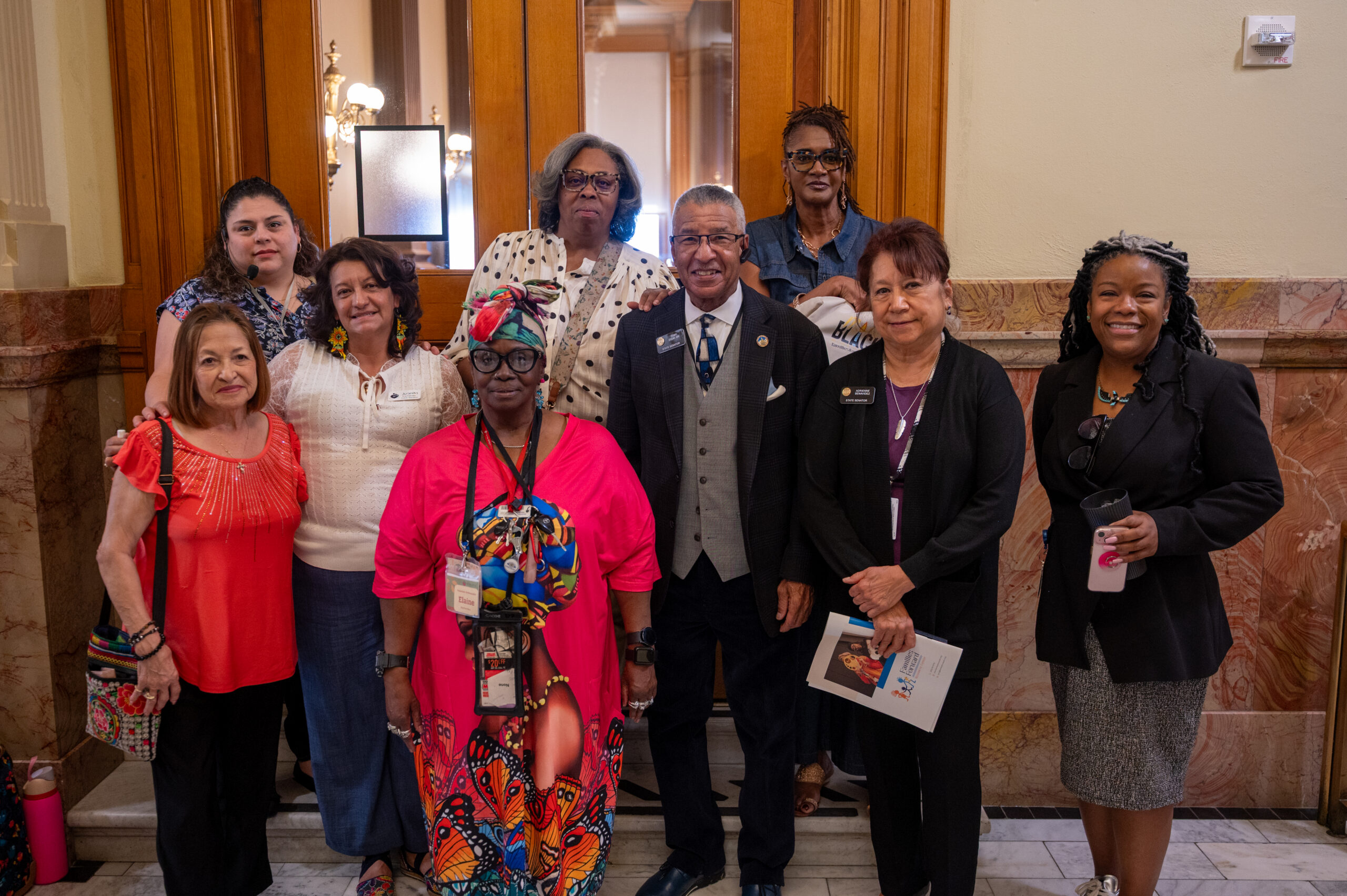 A group of Black and Latina women stand outside of wooden doors to the Senate committee chamber with Black and Latina Representatives and Senators wearing suits. 