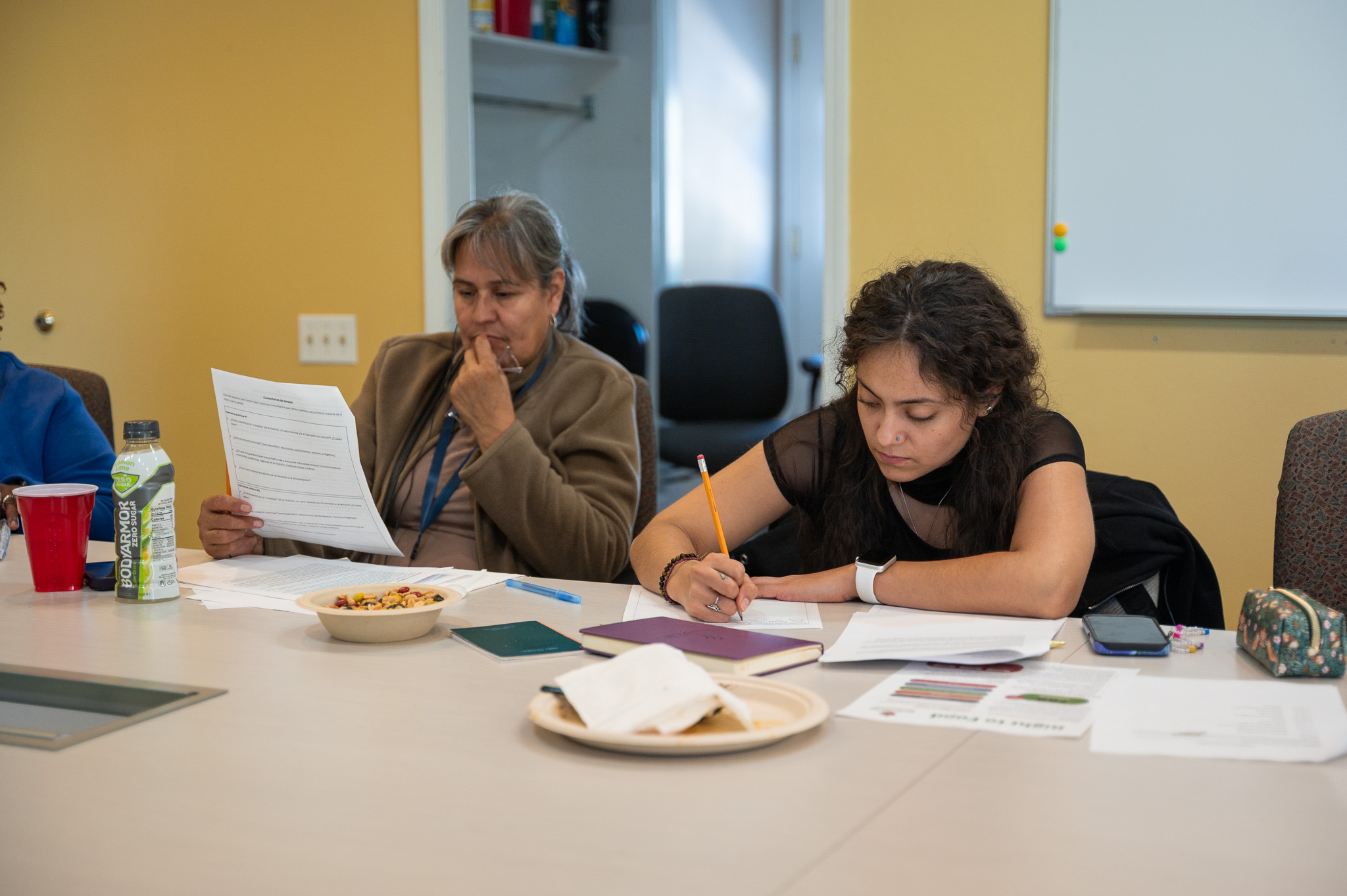 A young woman leans over the table writing on a piece of paper. An older woman sits next to her holding a paper, reading to herself.